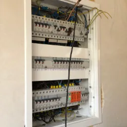 Otwarta skrzynka elektryczna podtynkowa z zainstalowanymi bezpiecznikami i przewodami, widoczne ślady prac wykończeniowych na ścianie wokół obudowy.