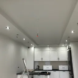 Wnętrze kuchni w trakcie montażu oświetlenia sufitowego typu spot, widoczna drabina i wiszące przewody elektryczne.