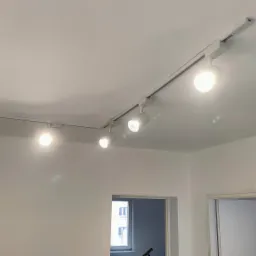 Biały sufit z zamontowanym systemem szynowym oświetleniowym z czterema reflektorami punktowymi, skierowanymi w różne strony, w nowym pomieszczeniu z widocznymi drzwiami i oknem.