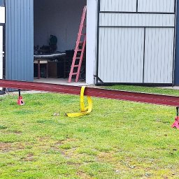 ZRKopytek - Czerwony profil stalowy podparty na podnośnikach na trawie, z żółtym pasem transportowym, na tle otwartych drzwi garażowych i warsztatu.