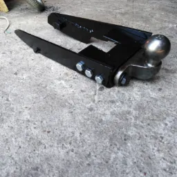 Czarny, metalowy hak holowniczy z kulą, przymocowany śrubami, leżący na szarej, betonowej posadzce w warsztacie.