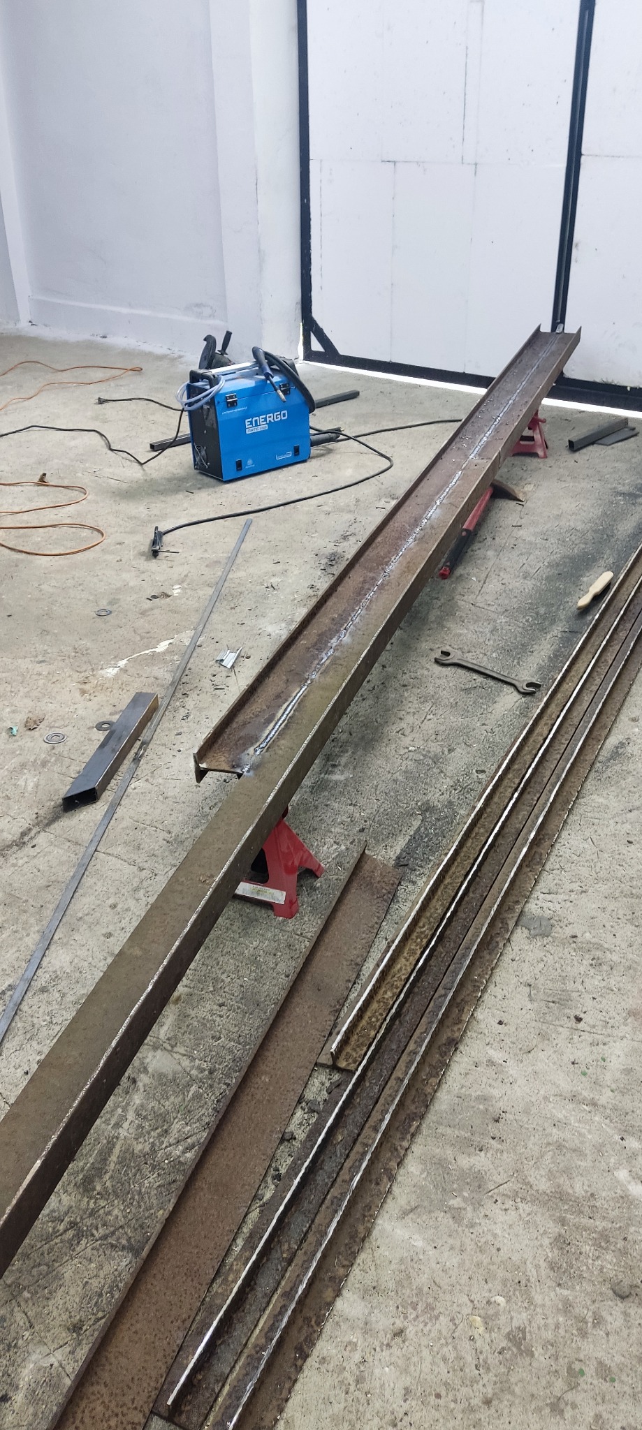 Spawany profil metalowy na kozłach spawalniczych, obok spawarka inwertorowa i narzędzia ślusarskie na betonowej posadzce.