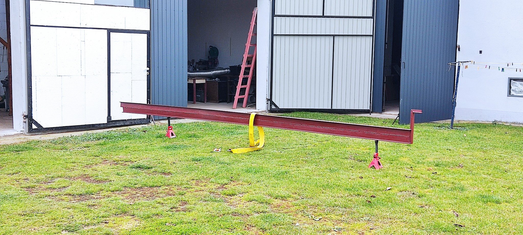 Czerwony profil stalowy podparty na podnośnikach na trawie, z żółtym pasem transportowym, na tle otwartych drzwi garażowych i warsztatu.