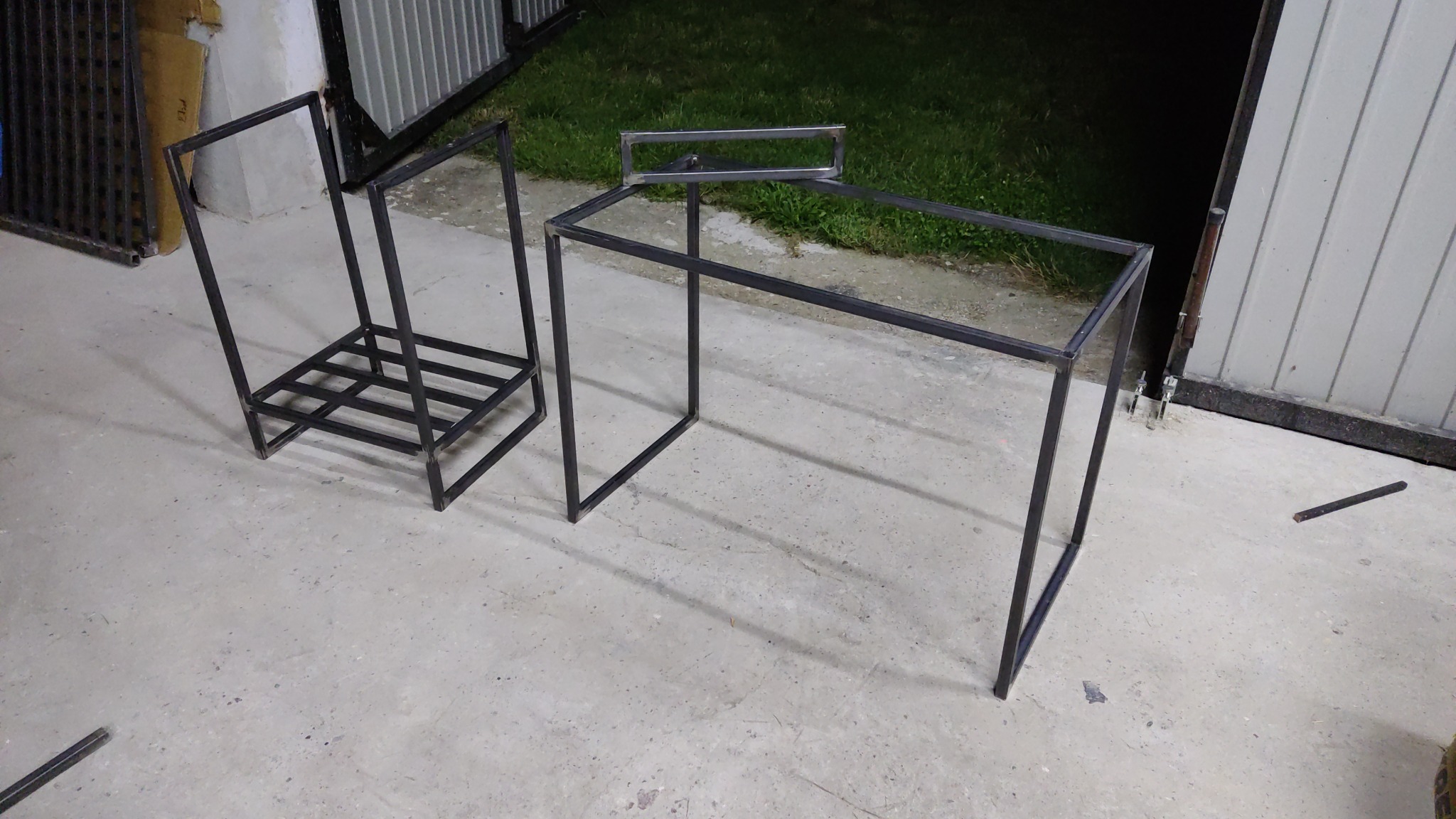 Konstrukcje metalowe: stelaż pod stolik kawowy i regał na drewno, spawane z profili stalowych, surowe wykończenie, przed garażem.