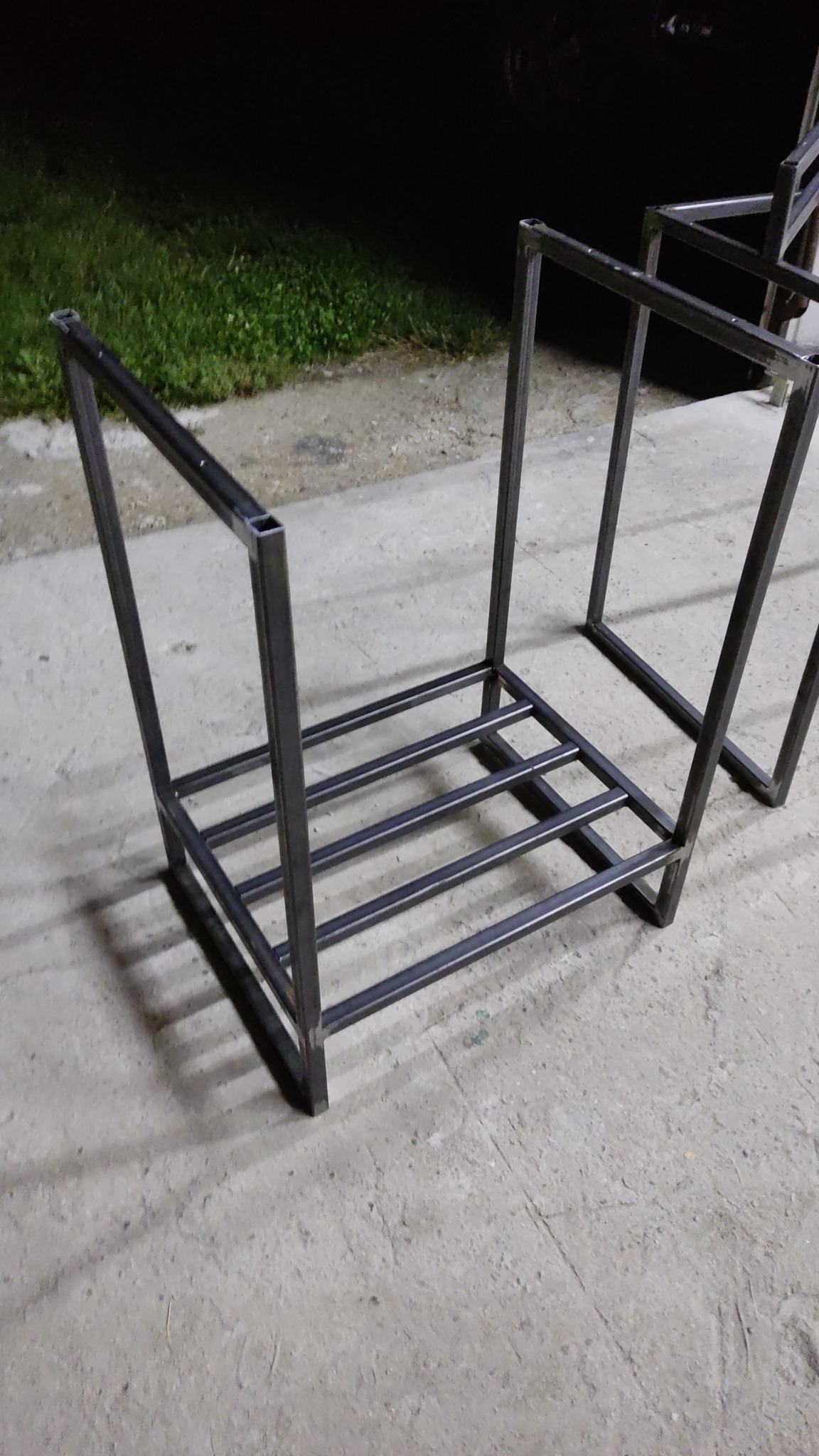 Surowa, spawana konstrukcja metalowa z profili kwadratowych, prawdopodobnie element większej całości, widoczna na betonowym podłożu w słabym oświetleniu.