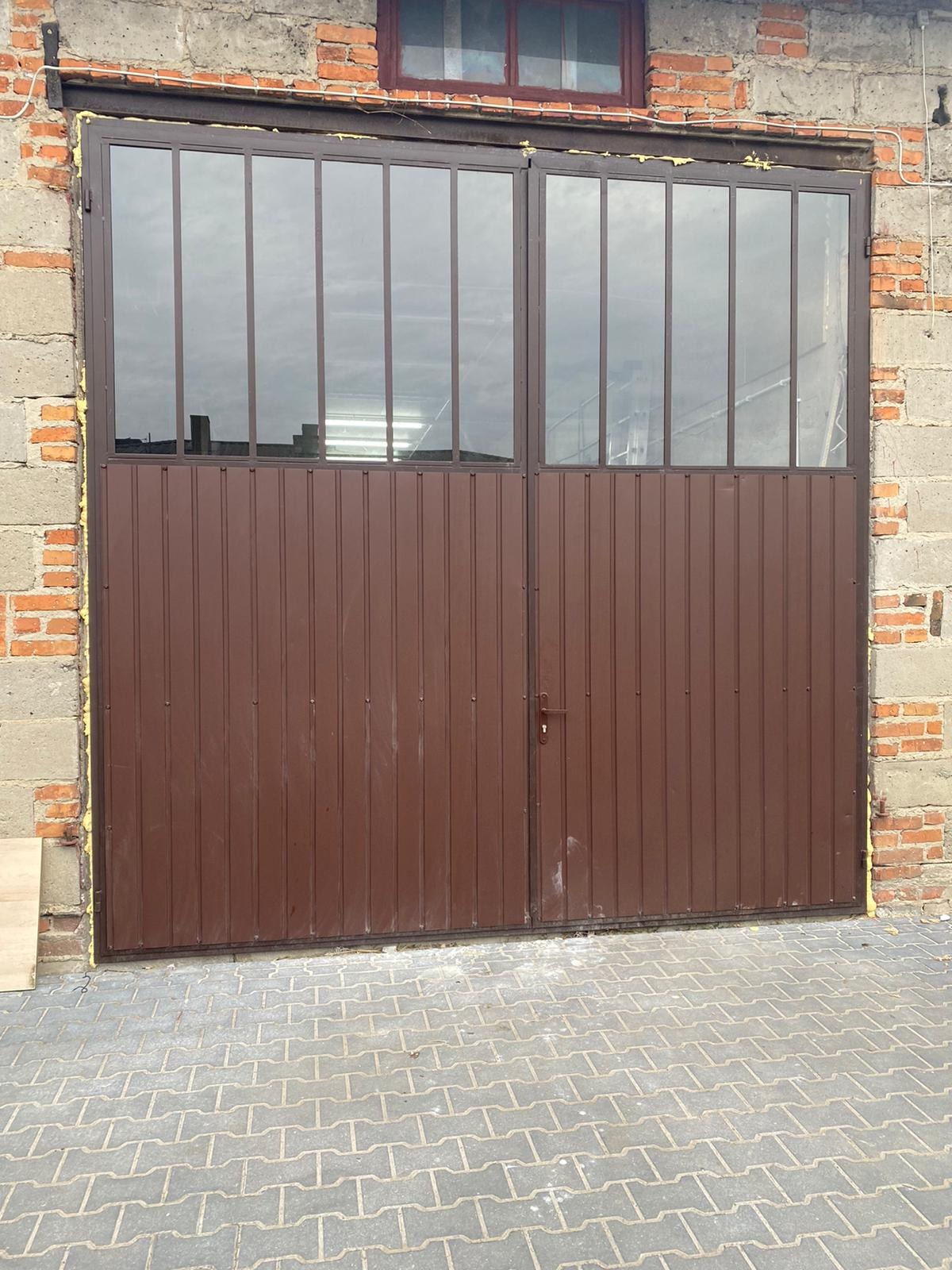 Brązowa brama przemysłowa z górną częścią oszkloną, zamontowana w surowej, ceglano-betonowej ścianie budynku z widocznym oknem na górze i kostką brukową przed bramą.