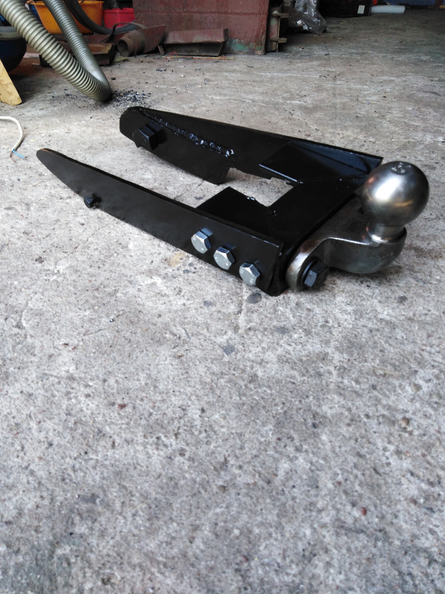 Czarny, metalowy hak holowniczy z kulą, przymocowany śrubami, leżący na szarej, betonowej posadzce w warsztacie.