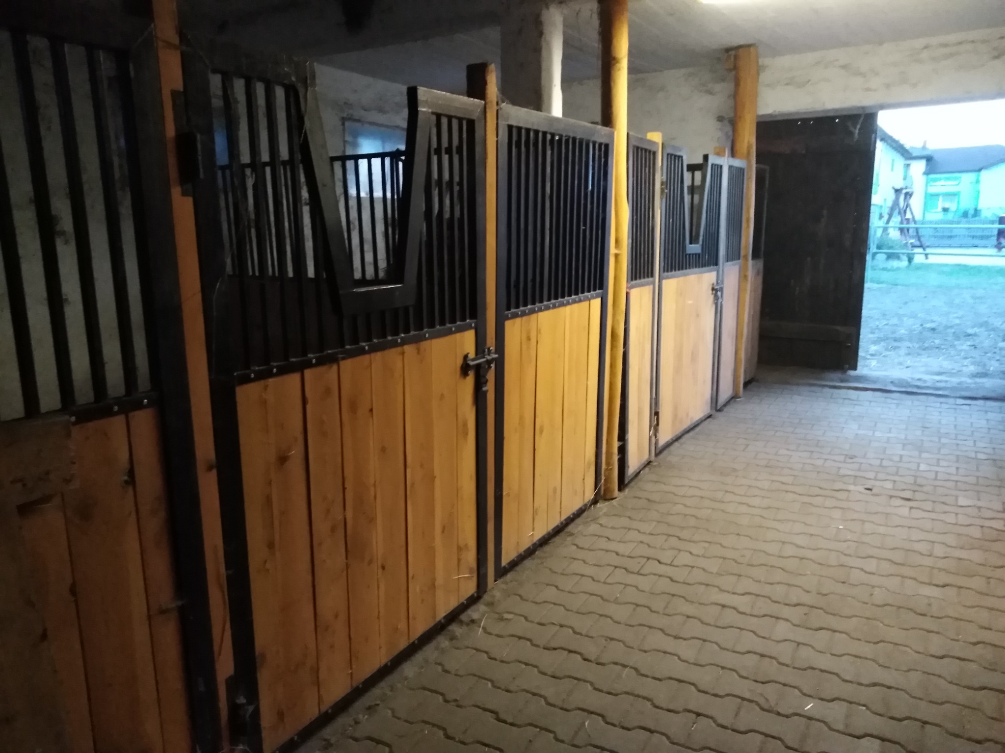 Wnętrze stajni z rzędem drewniano-metalowych boksów dla koni, z widocznym zamknięciem na jeden z nich i wyjściem na zewnątrz.