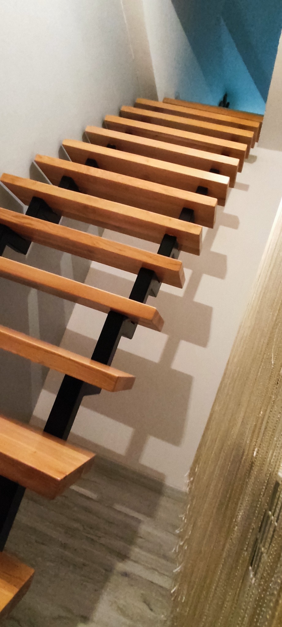 Ażurowe schody z drewnianymi stopniami i czarną metalową konstrukcją nośną, widziane z dołu pod kątem, z widocznymi cieniami na ścianie i podłodze.