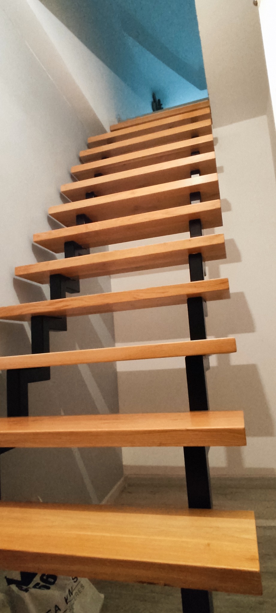 Nowoczesne schody z drewnianymi stopniami i czarnymi metalowymi wspornikami, widok z dołu, jasne tło ścian i sufitu.