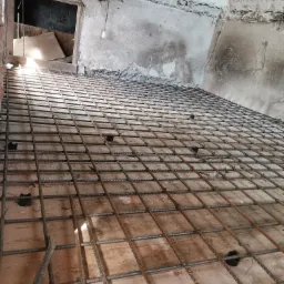 Przygotowanie zbrojenia pod wylewkę betonową na nierównym, starym podłożu wewnątrz remontowanego pomieszczenia z widocznym otworem drzwiowym i wiszącą żarówką.