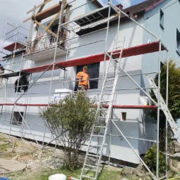 Pracownik na rusztowaniu z wiertarką, podczas prac wykończeniowych elewacji domu jednorodzinnego, z widocznymi śladami po niedawno położonym tynku.