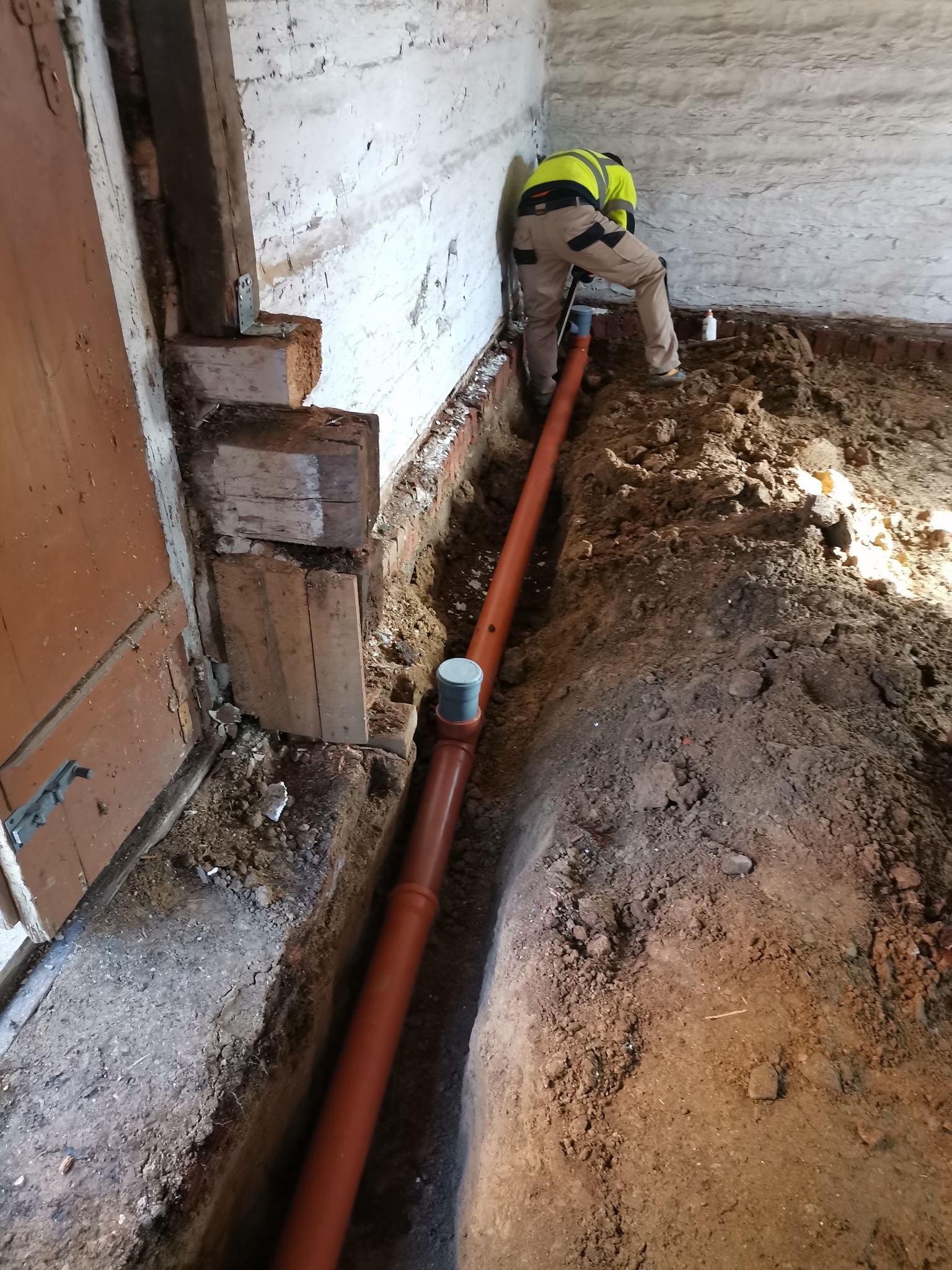 Instalacja rury kanalizacyjnej w wykopie wewnątrz budynku z ceglaną ścianą, pracownik w odblaskowej kamizelce w trakcie pracy.