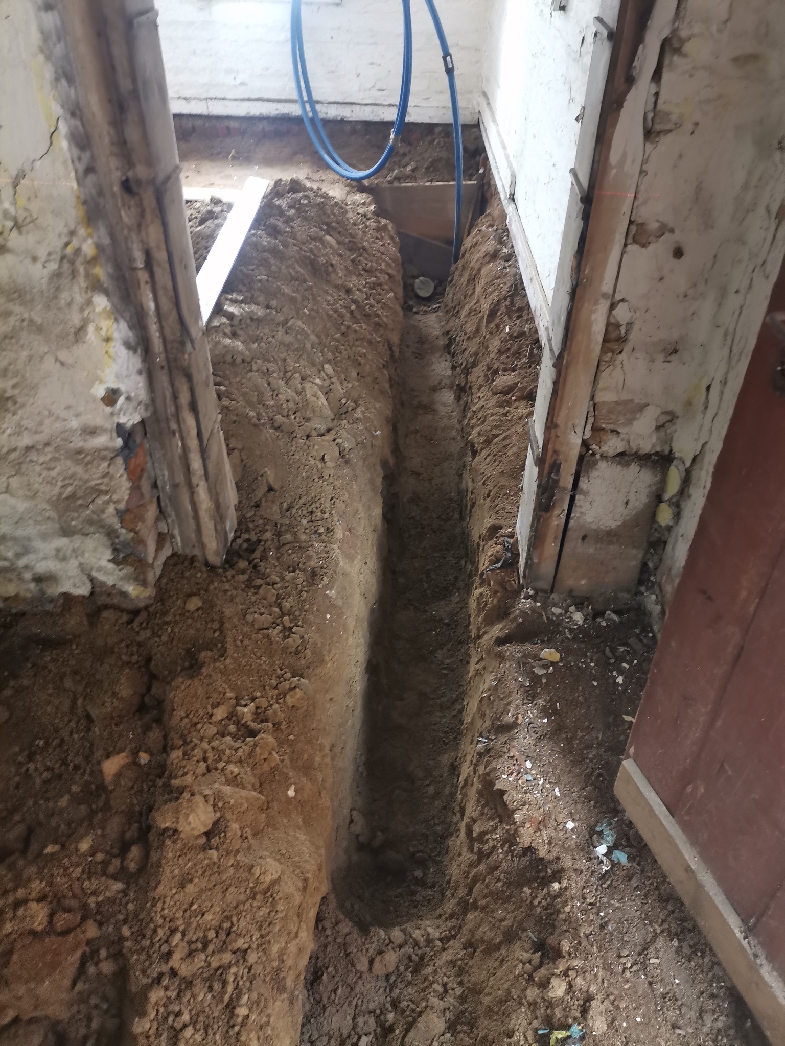 Wykop w ziemi wewnątrz budynku z widocznymi instalacjami hydraulicznymi i fragmentami ścian w stanie surowym.