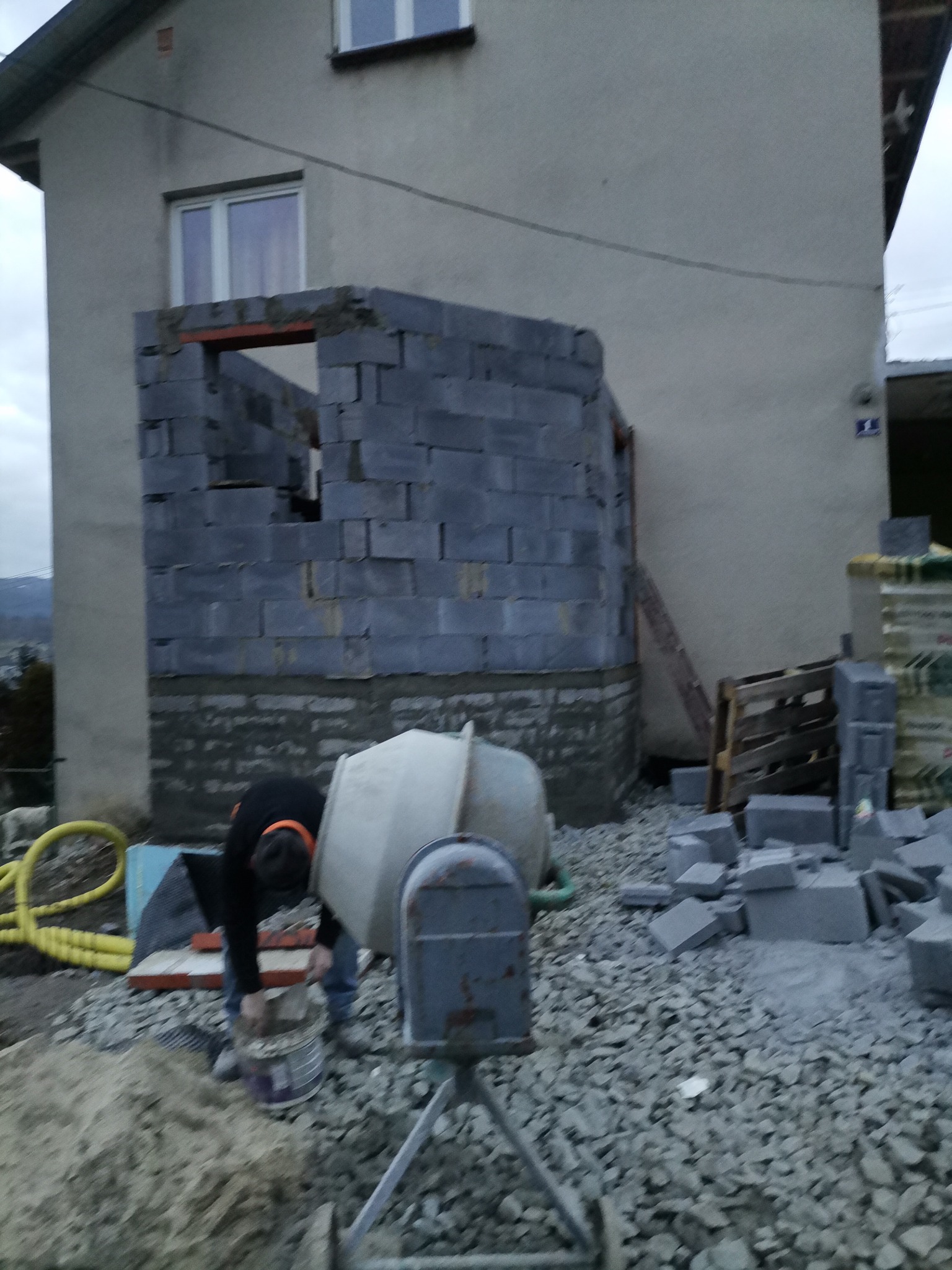 Wznoszenie ściany z szarych bloczków betonowych przy pomocy betoniarki i zaprawy, widoczny pracownik w trakcie nabierania zaprawy z wiadra, otoczenie budowy z gruzem i piaskiem.