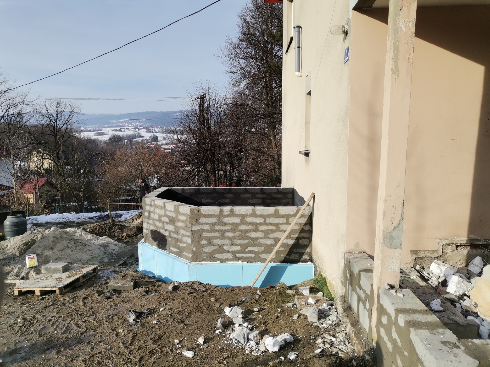 Wznoszenie ścian z bloczków betonowych na fundamencie zabezpieczonym niebieskim styropianem, widoczny fragment elewacji budynku i sterta piasku.