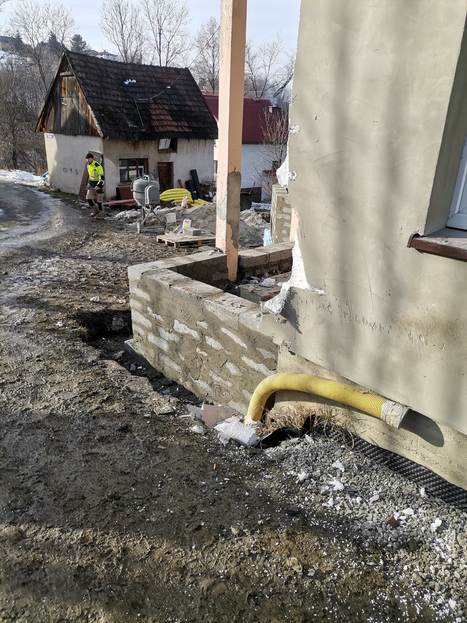 Budowa ściany z bloczków betonowych przy otynkowanej elewacji domu, z widocznym żółtym wężem wentylacyjnym i pracownikiem w odblaskowej kamizelce w tle.