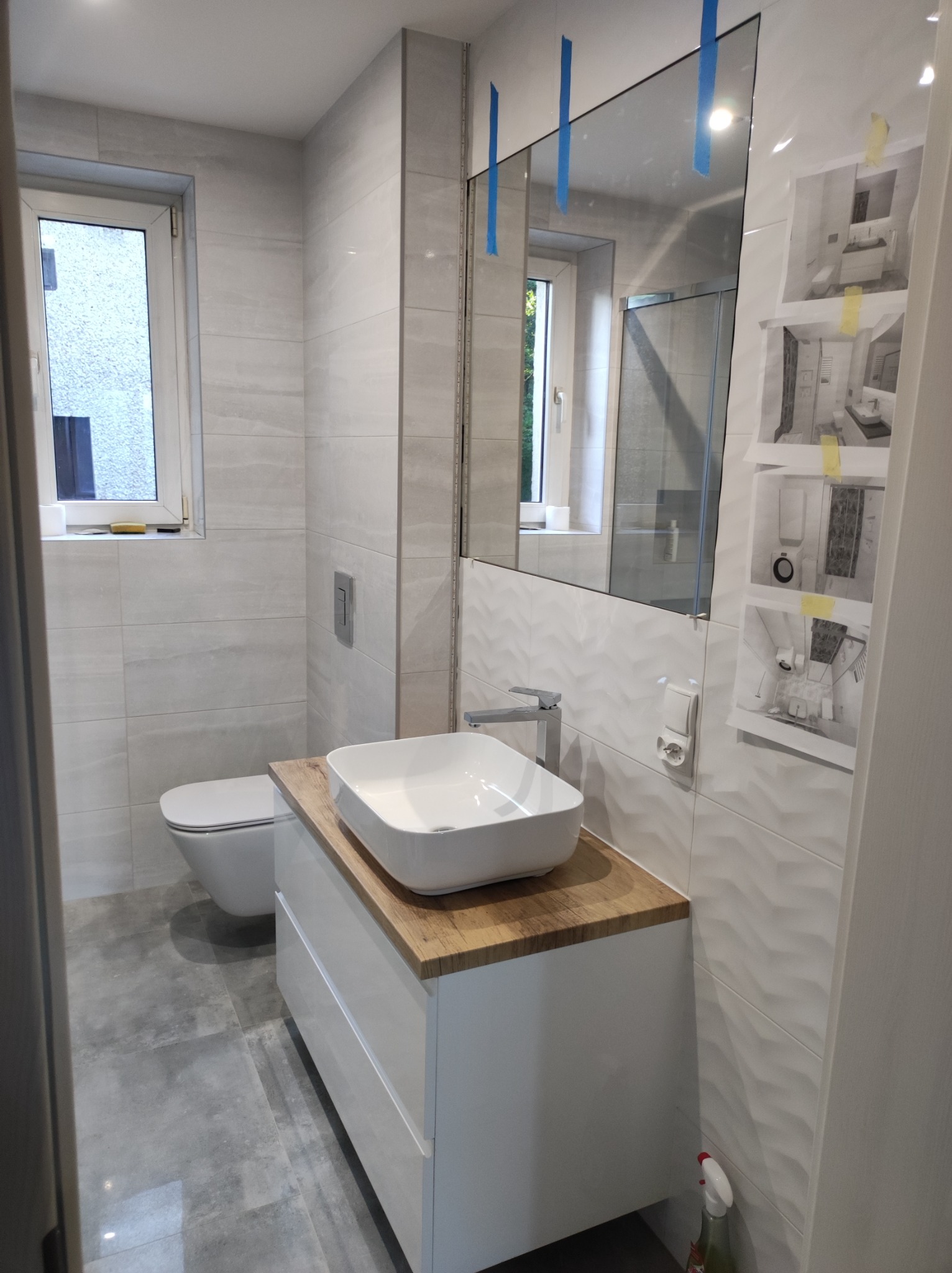 Nowoczesna łazienka w szaro-białej kolorystyce z drewnianym blatem pod umywalką, widoczne płytki imitujące kamień, lustro z niebieską taśmą zabezpieczającą i projekt łazienki na ścianie.