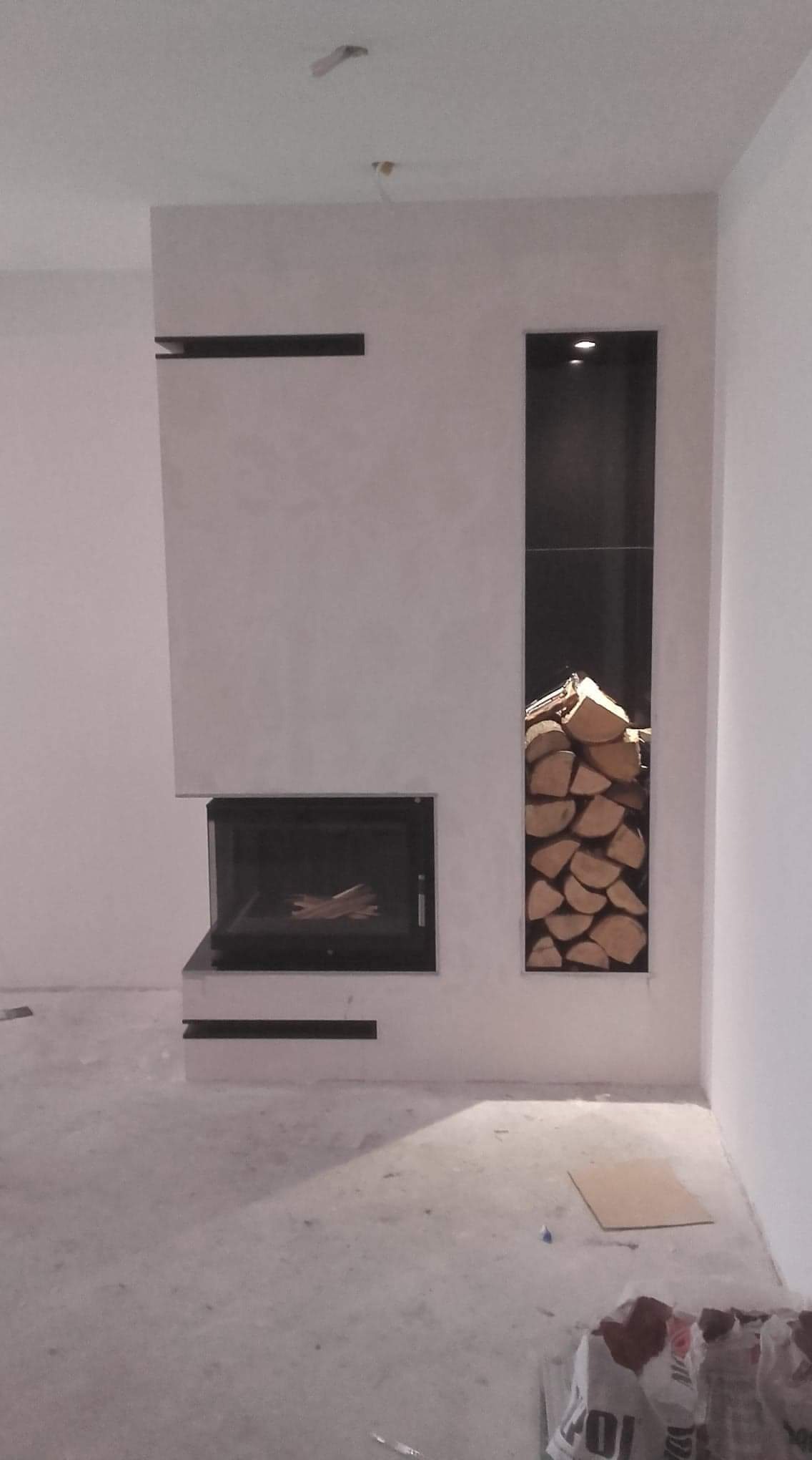 Nowoczesny kominek z wkładem kominkowym i wnęką na drewno, wykończony szarym tynkiem strukturalnym, widok na surową podłogę w trakcie prac wykończeniowych.