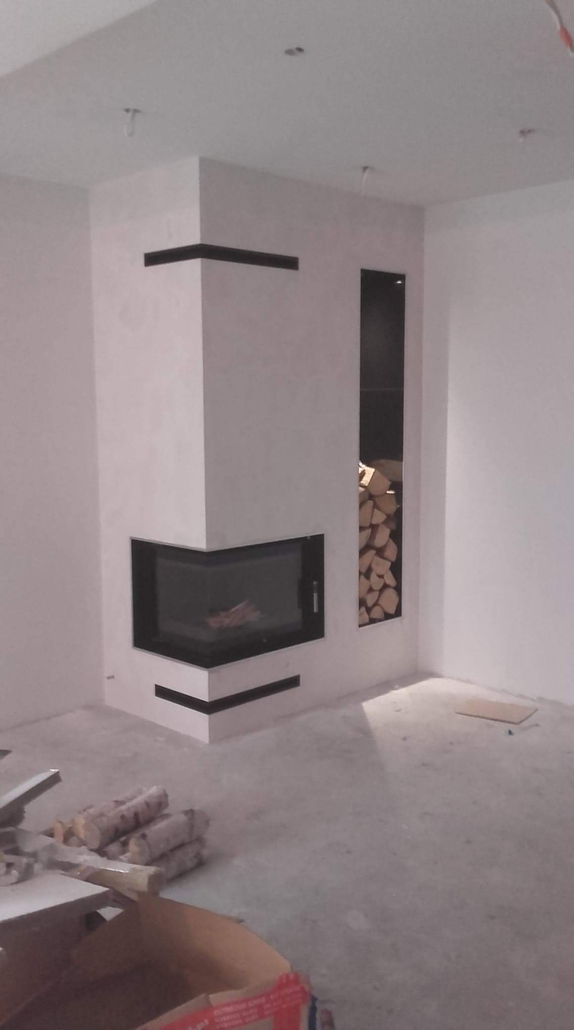 Nowoczesny kominek narożny z czarnym obramowaniem i wnęką na drewno, surowy beton na ścianach, podłoga w trakcie wykończenia, widoczne drewno do kominka, minimalistyczny design.