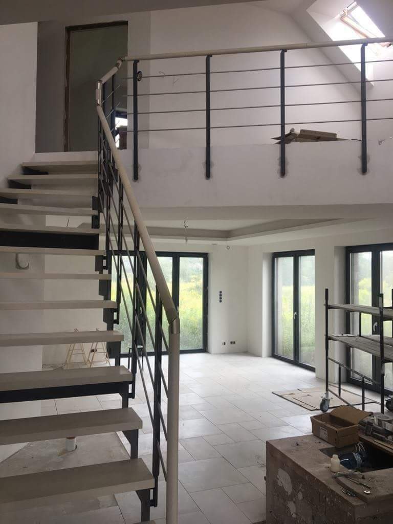 Wnętrze domu w trakcie wykańczania: schody z metalową balustradą, jasne płytki podłogowe, duże okna z widokiem na zieleń, widoczne materiały budowlane i rusztowanie.