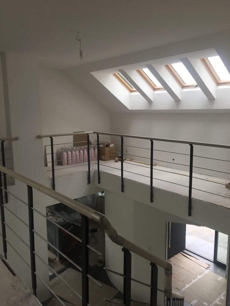 Wnętrze domu w trakcie wykończenia: widok na schody z metalową balustradą, jasne ściany, okna dachowe i materiały budowlane składowane na piętrze.