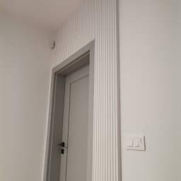 DOM ODNOWA DARIUSZ MARKIEWICZ - Nowoczesne wykończenie ściany z dekoracyjnymi listwami pionowymi wokół szarej ościeżnicy i białych drzwi, widoczny włącznik światła na ścianie.