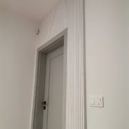 Nowoczesne wykończenie ściany z dekoracyjnymi listwami pionowymi wokół szarej ościeżnicy i białych drzwi, widoczny włącznik światła na ścianie.
