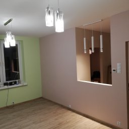 DOM ODNOWA DARIUSZ MARKIEWICZ - Pomalowane na różowo ściany, drewniana podłoga, okno z zieloną ramą, trzy lampy wiszące z wzorzystymi kloszami i widok na sąsiednie pomieszczenie przez otwór w ścianie z trzema cylindrycznymi lampami.