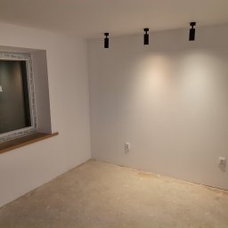 DOM ODNOWA DARIUSZ MARKIEWICZ - Wnętrze pomieszczenia w trakcie prac wykończeniowych: surowy beton na podłodze, białe ściany, czarne reflektory sufitowe, okno z drewnianym parapetem, widoczne gniazdka elektryczne.