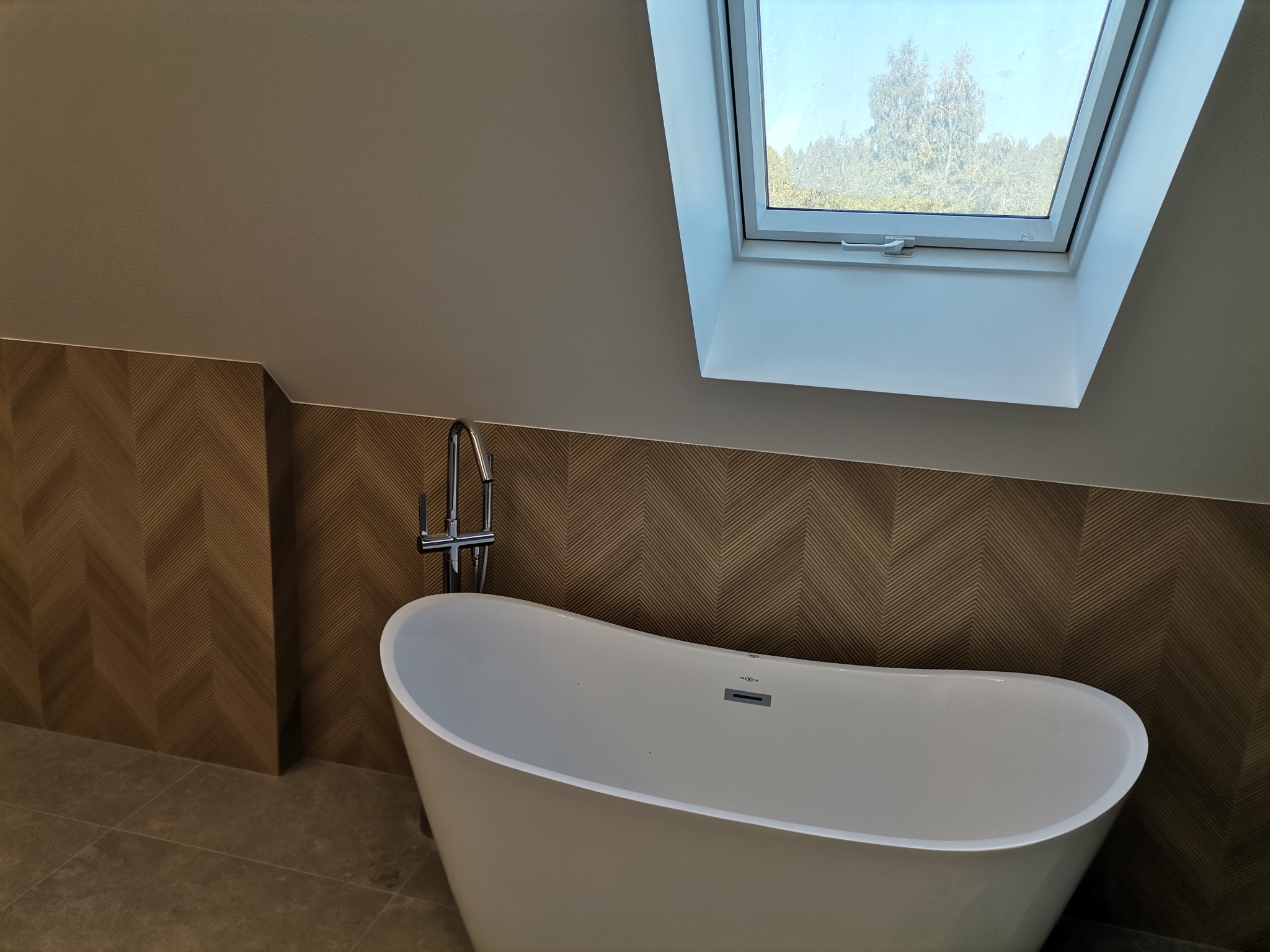 Nowoczesna biała wanna wolnostojąca z chromowaną armaturą na tle ściany wyłożonej płytkami w jodełkę, oświetlona światłem wpadającym przez okno dachowe.
