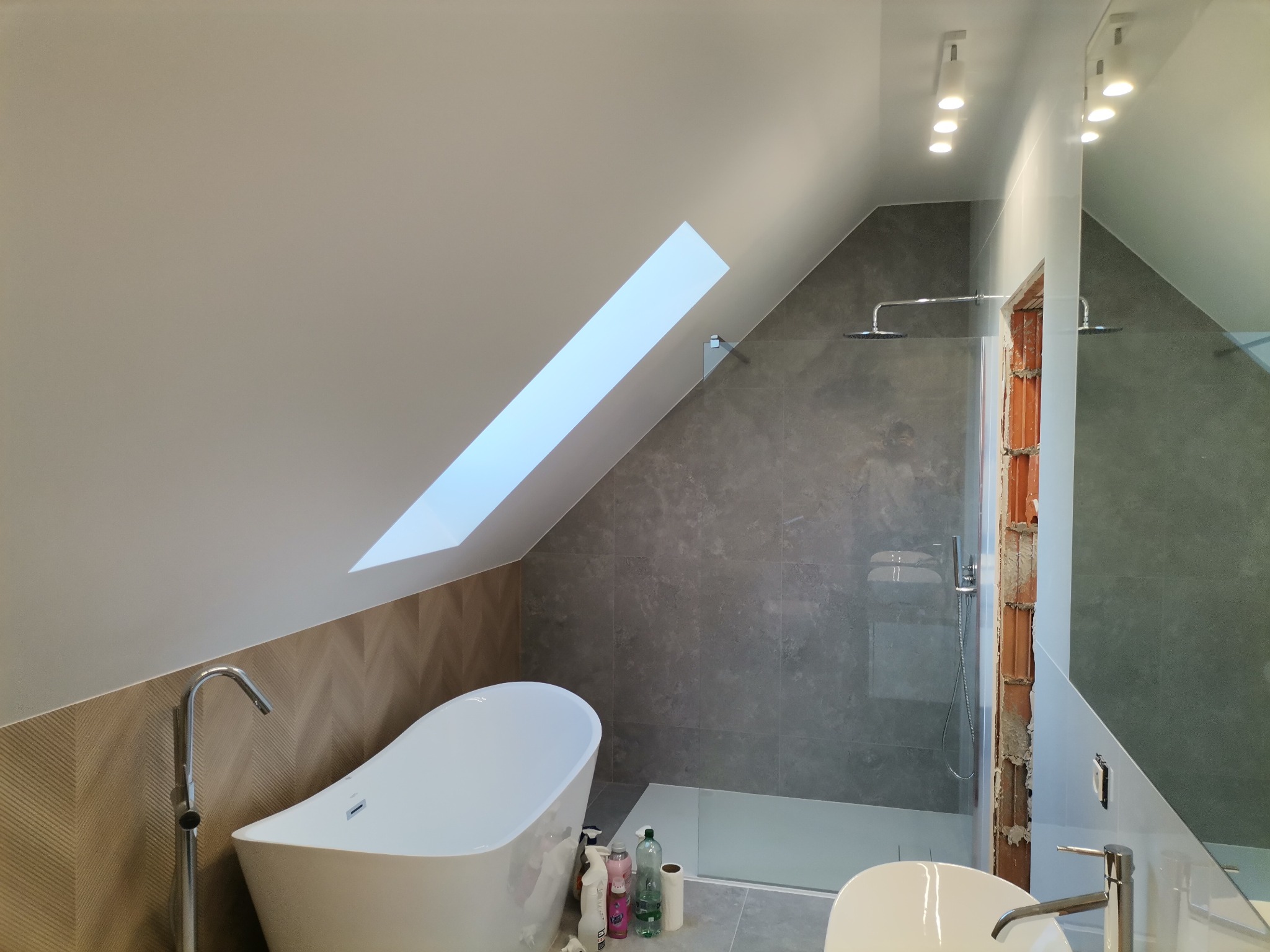 Nowoczesna łazienka na poddaszu z oknem dachowym, wolnostojąca wanna obok prysznica typu walk-in z szarymi płytkami imitującymi beton, widoczny fragment niezakończonego wykończenia przy lustrze.