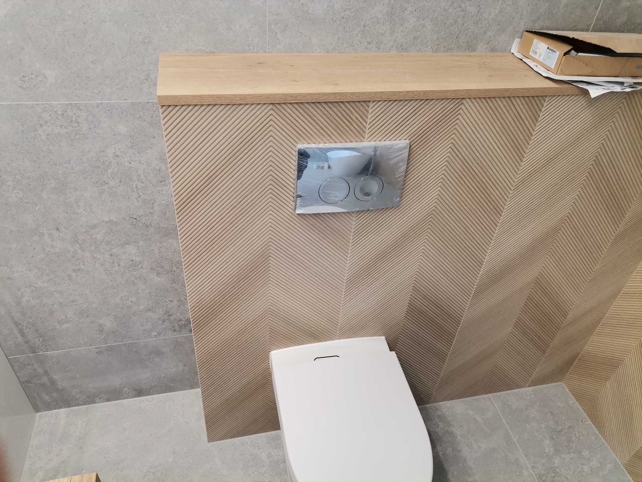 Podwieszana toaleta z białą deską, na tle ściany wyłożonej płytkami z wzorem jodełki w odcieniach beżu, z zabudową z drewnianym blatem, na którym leży karton z akcesoriami.