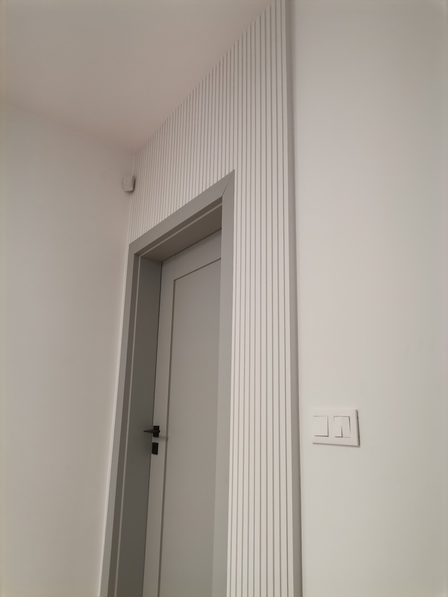 Nowoczesne wykończenie ściany z dekoracyjnymi listwami pionowymi wokół szarej ościeżnicy i białych drzwi, widoczny włącznik światła na ścianie.