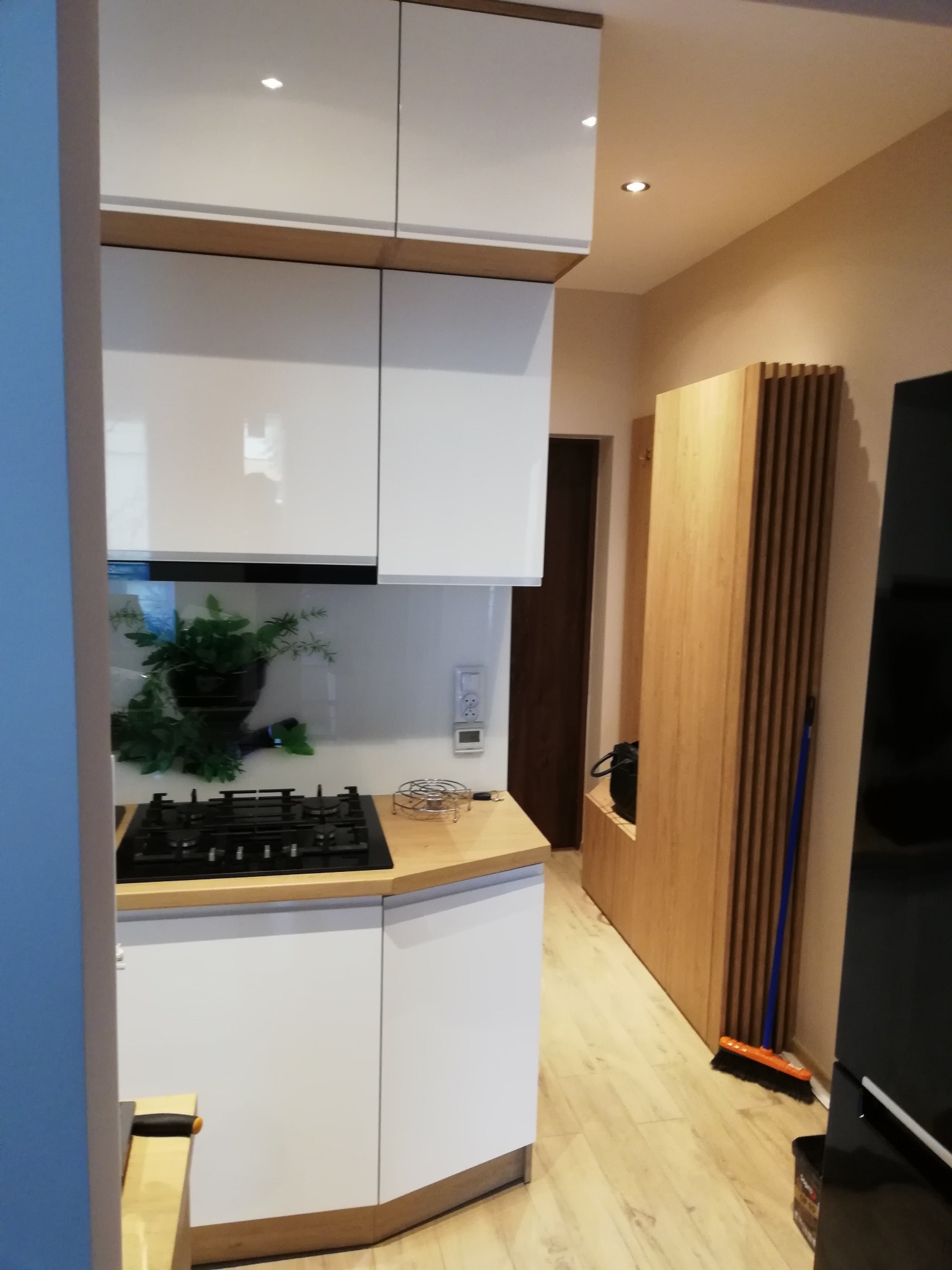 Nowoczesna kuchnia z białymi szafkami i drewnianymi akcentami, widoczna płyta gazowa, rośliny w doniczce na blacie, drewniana zabudowa ściany z wnęką i miotłą opartą o ścianę.