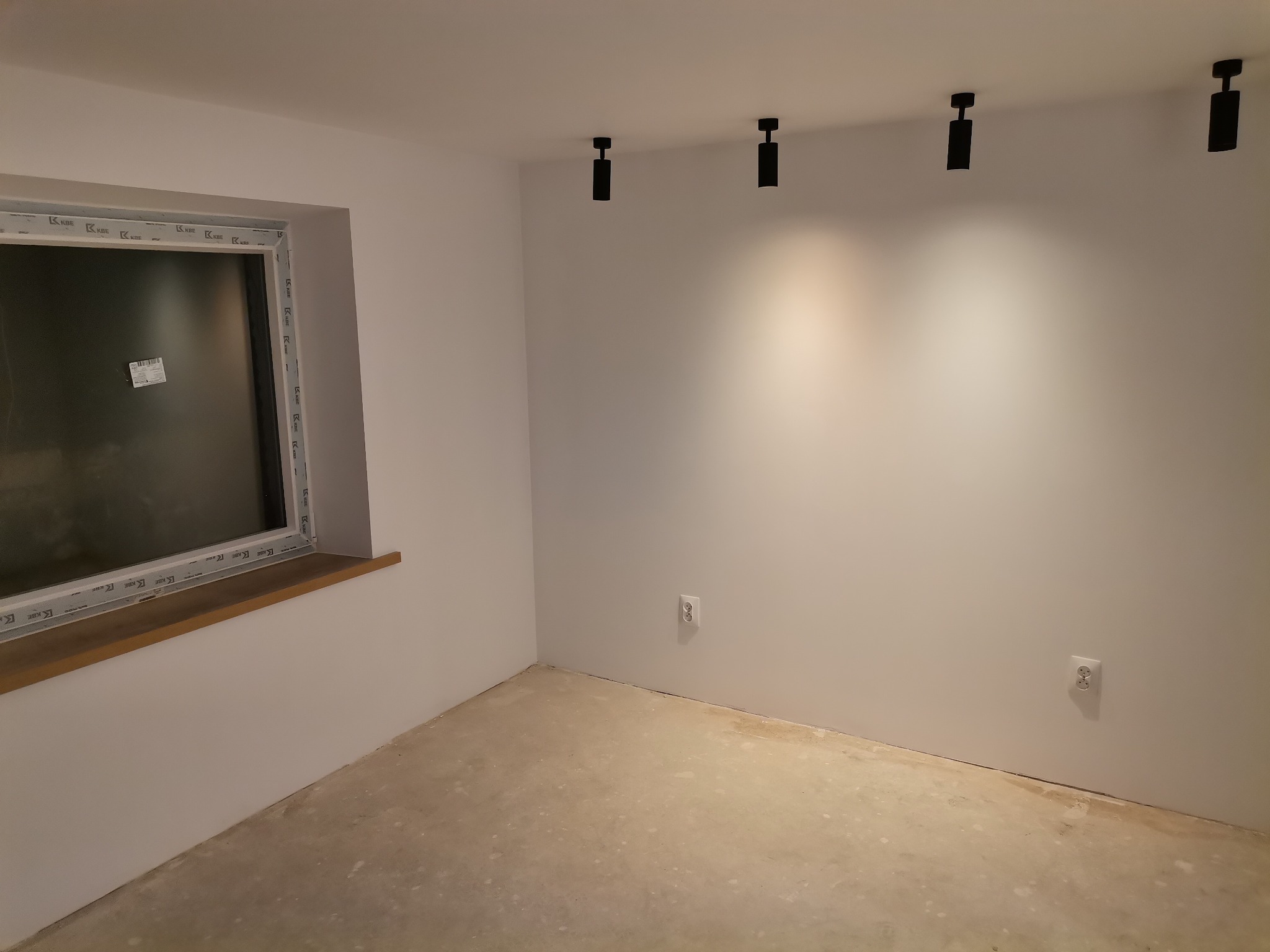 Wnętrze pomieszczenia w trakcie prac wykończeniowych: surowy beton na podłodze, białe ściany, czarne reflektory sufitowe, okno z drewnianym parapetem, widoczne gniazdka elektryczne.