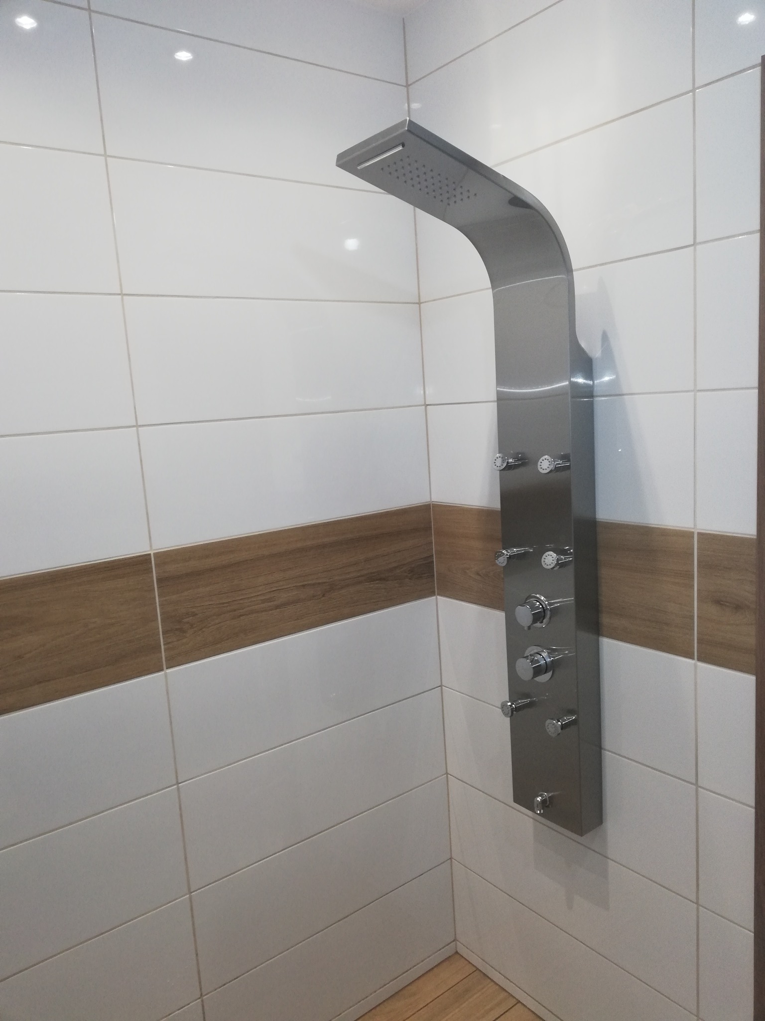 Nowoczesna kolumna prysznicowa ze stali nierdzewnej z wieloma dyszami, zamontowana na ścianie wyłożonej białymi płytkami z poziomym pasem dekoracyjnym imitującym drewno w wyremontowanej łazience.