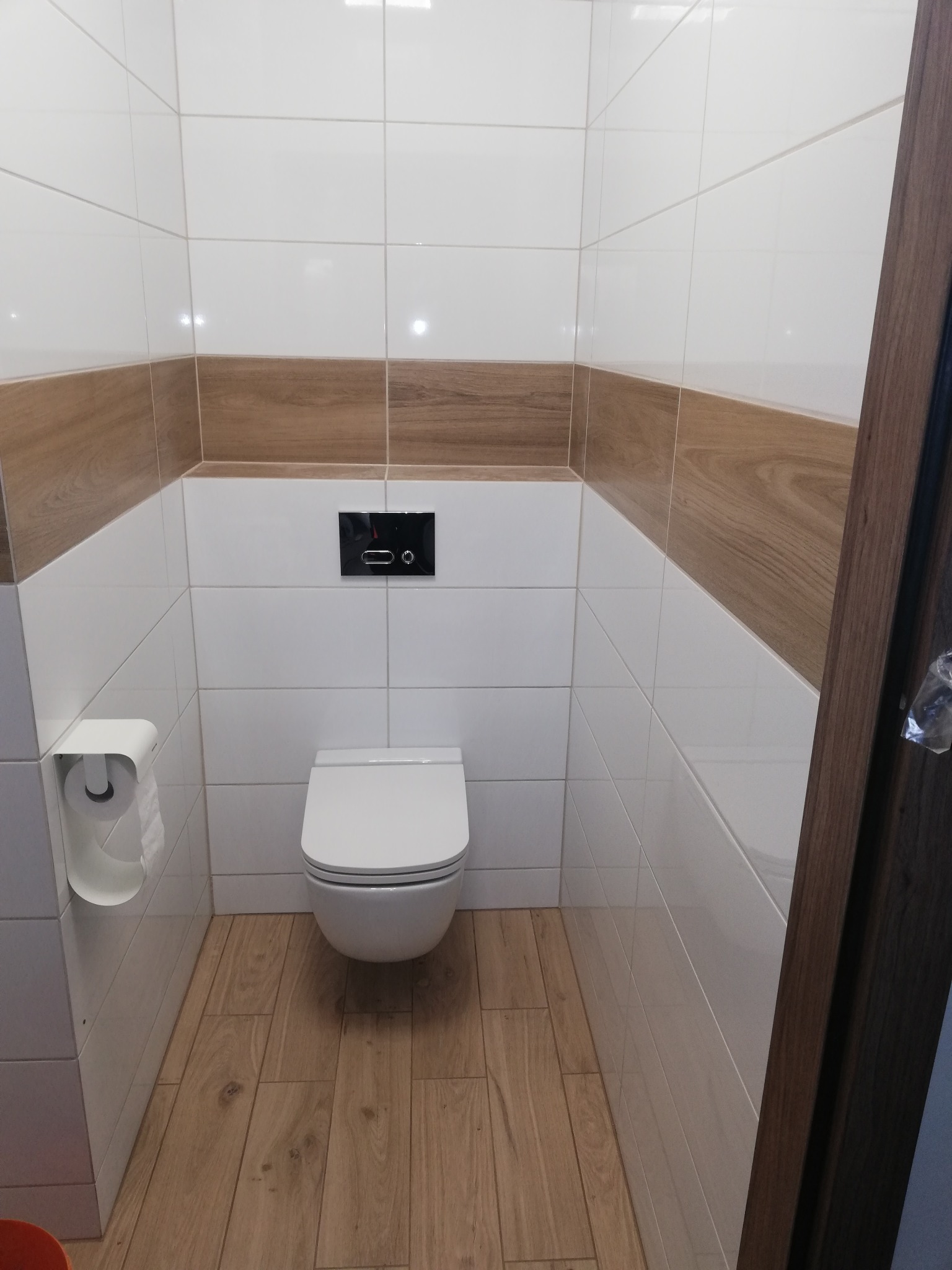 Nowoczesna toaleta podwieszana w wykończonej łazience, białe płytki z drewnopodobnym pasem, drewniana podłoga, czarny przycisk spłukujący.