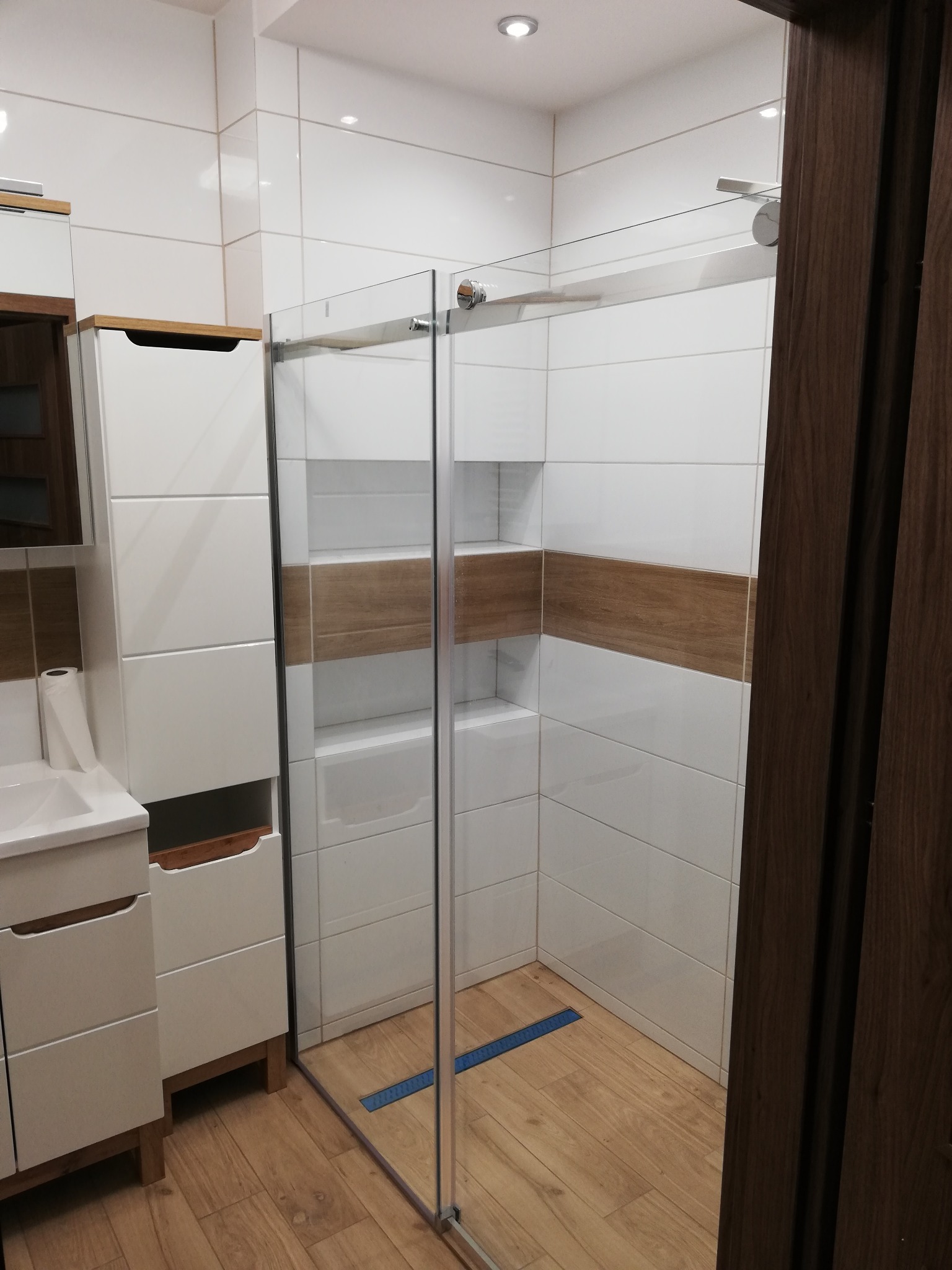 Nowoczesna łazienka z kabiną prysznicową typu walk-in, białe płytki z drewnopodobnym pasem, drewniana podłoga i szafka pod umywalką z drewnianymi elementami.