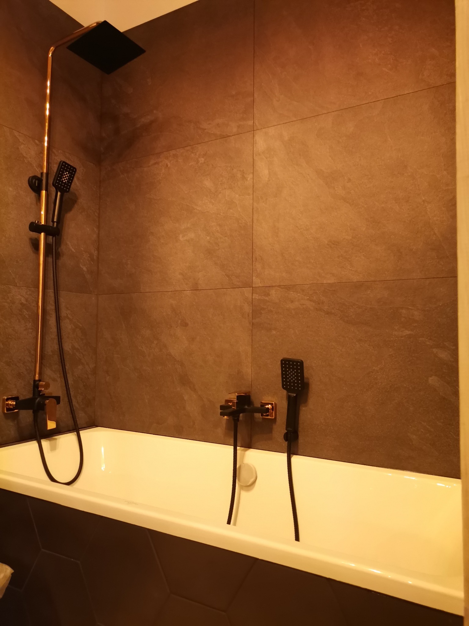 Nowoczesna łazienka z wanną, armaturą w kolorze złota i czerni, oraz brązowymi płytkami imitującymi kamień na ścianie. Widoczny zestaw prysznicowy z deszczownicą i słuchawką, bateria wannowa,...