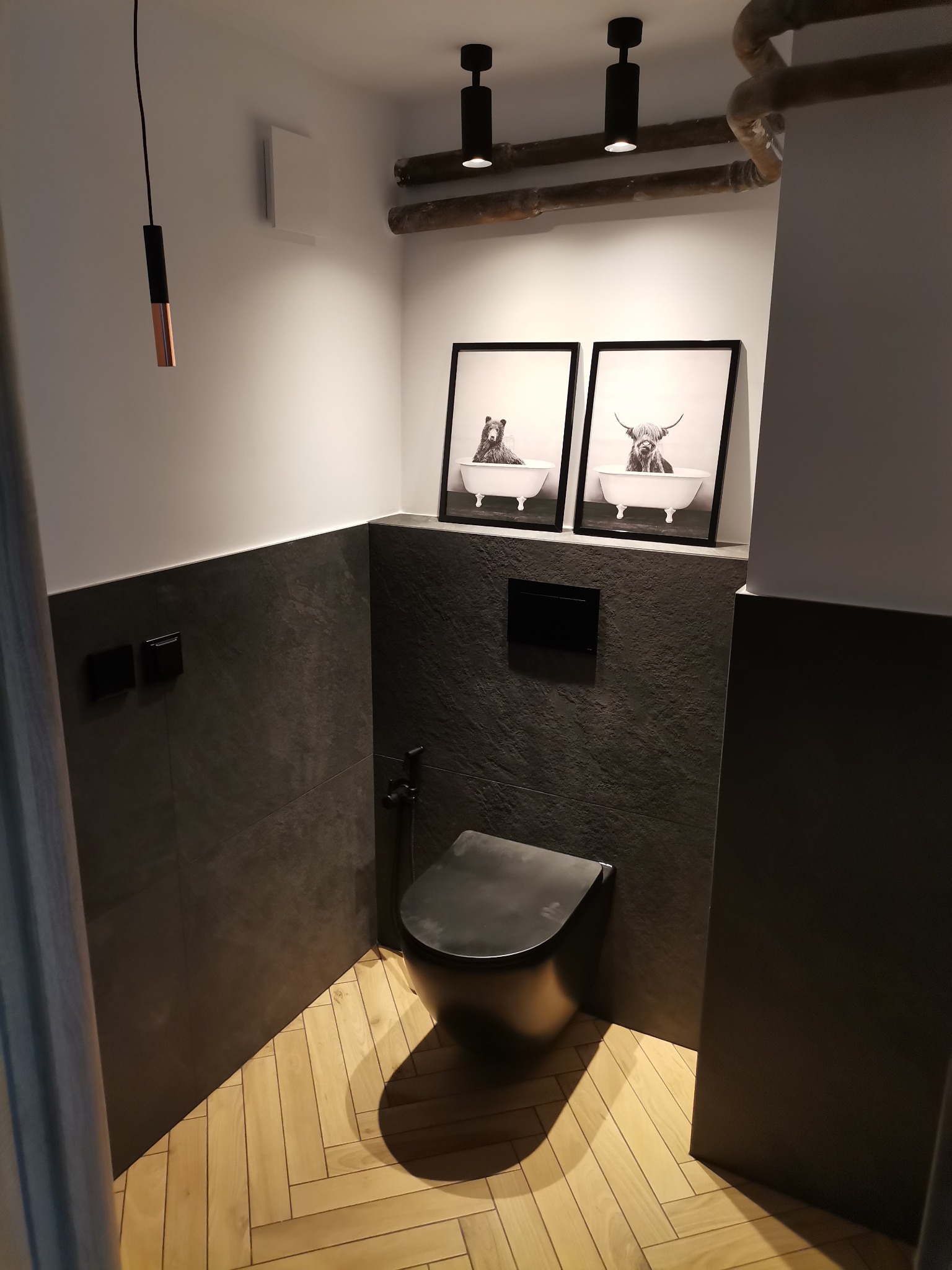 Nowoczesna toaleta w łazience z szarymi płytkami imitującymi kamień, drewnianą podłogą ułożoną w jodełkę i czarną wiszącą miską WC. Na ścianie wiszą czarno-białe grafiki zwierząt w wannie. Widoczne...