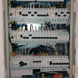 Otwarta skrzynka rozdzielcza z licznymi bezpiecznikami, przewodami i listwami zaciskowymi, zamontowana w ścianie z widocznymi śladami niedokończonego wykończenia.