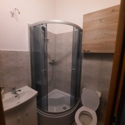 Artur Biernacki - Niewielka łazienka z narożną kabiną prysznicową z grafitowymi profilami, umywalką z szafką, wiszącą szafką w kolorze drewna i otwartą toaletą. Na ścianie widoczne wystające kable.