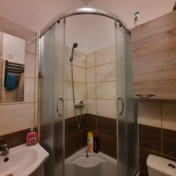 Artur Biernacki - Narożna kabina prysznicowa z matowego szkła, beżowo-brązowe płytki, umywalka z kosmetykami, wisząca szafka i toaleta w małej łazience.