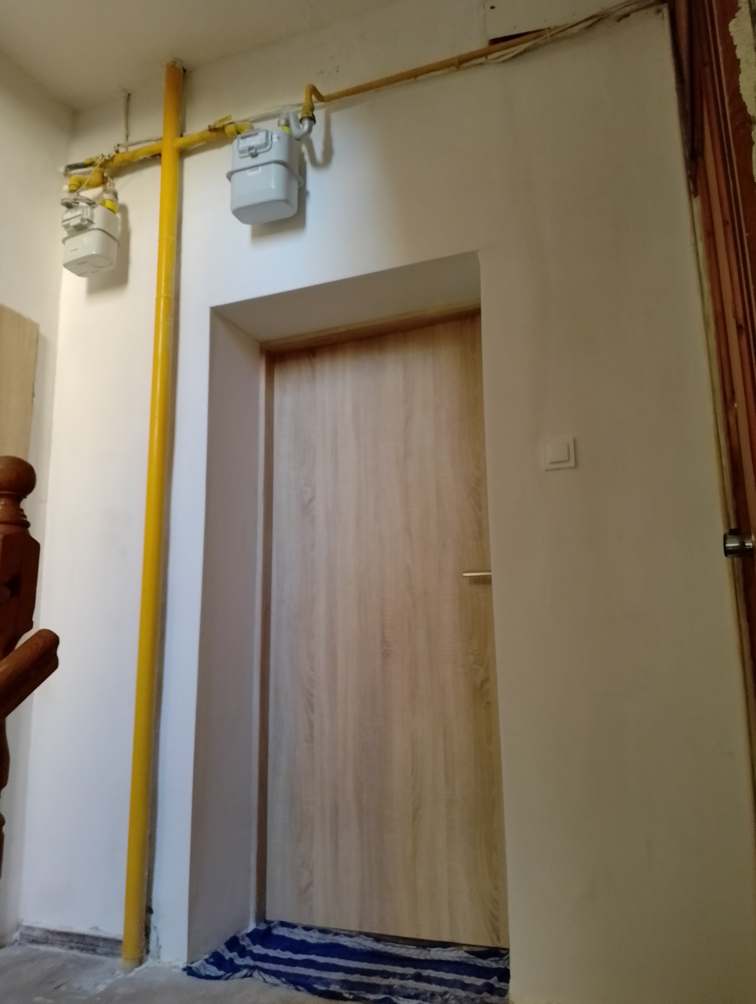 Zamontowane drzwi wewnętrzne o jasnym, drewnopodobnym wykończeniu, widoczne na tle białej ściany z żółtą rurą gazową i licznikiem, na podłodze leży niebieska szmata.