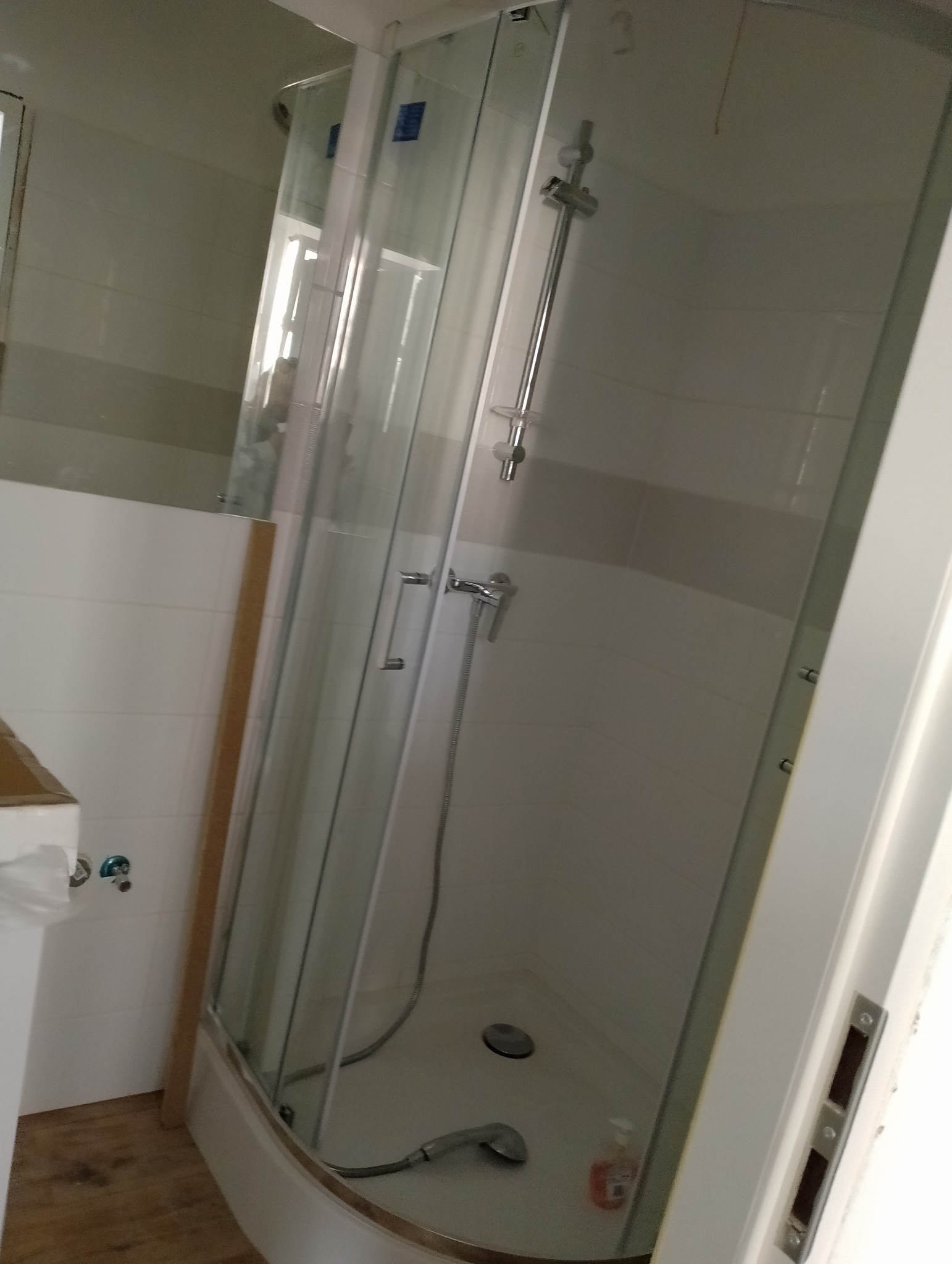 Kabina prysznicowa z chromowaną armaturą, białe płytki z szarym pasem, widoczna butelka z płynem do mycia na dnie brodzika.