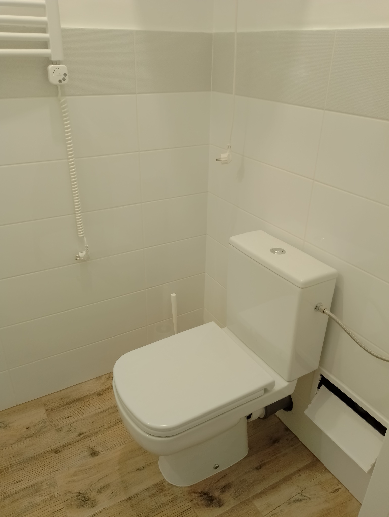 Nowoczesna toaleta kompaktowa w jasnej łazience z drewnopodobną podłogą i białymi płytkami, widoczny grzejnik drabinkowy z termostatem.