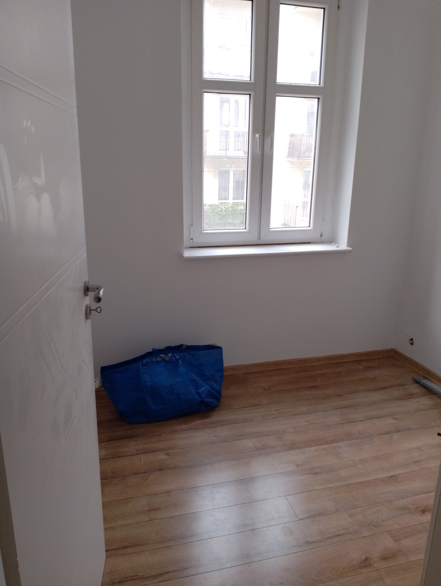 Pusty pokój po remoncie z nową podłogą z paneli, widok na okno i niebieska torba na podłodze.