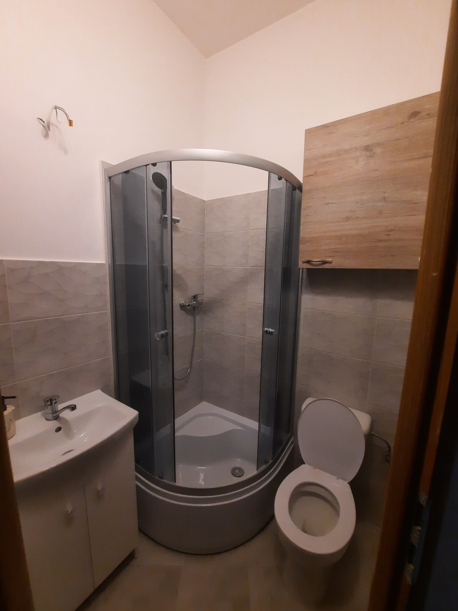 Niewielka łazienka z narożną kabiną prysznicową z grafitowymi profilami, umywalką z szafką, wiszącą szafką w kolorze drewna i otwartą toaletą. Na ścianie widoczne wystające kable.