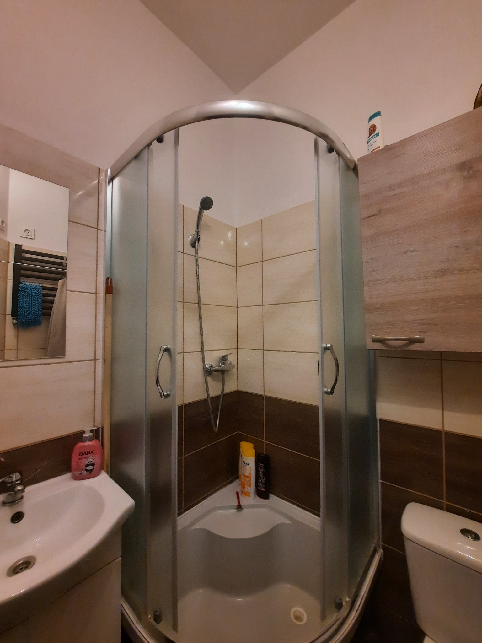 Narożna kabina prysznicowa z matowego szkła, beżowo-brązowe płytki, umywalka z kosmetykami, wisząca szafka i toaleta w małej łazience.